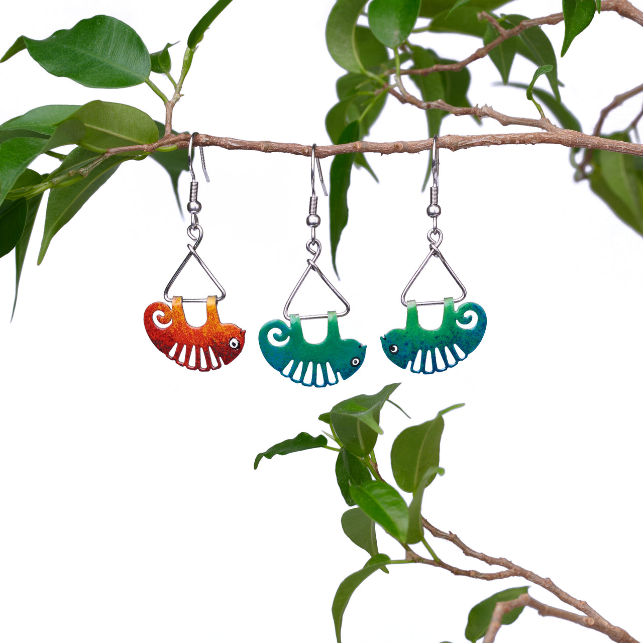 chameleon earrings on are hanging on a twig. One chameleon is red and there are two green chameleons